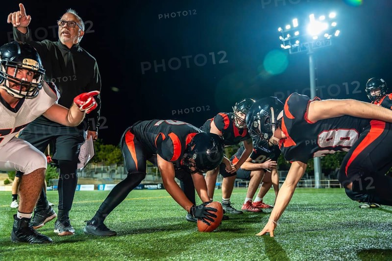 American football team huddled on a dark stadium field under bright lights at night, coach pointing and instructing players as they prepare for the next play and strategy