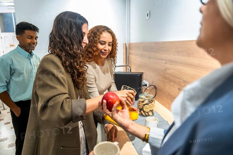 Coworkers gathering around a counter, offering an apple and orange juice, fostering team building and collaboration in a diverse and inclusive office environment during a light snack break