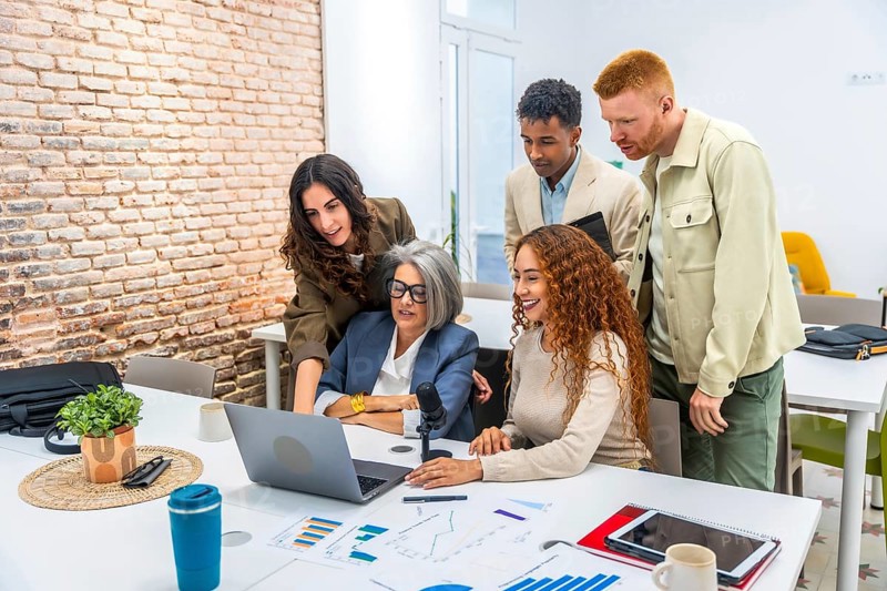 Diverse business professionals working together around a laptop, analyzing financial charts and presenting ideas during a casual office meeting, showcasing teamwork and modern communication