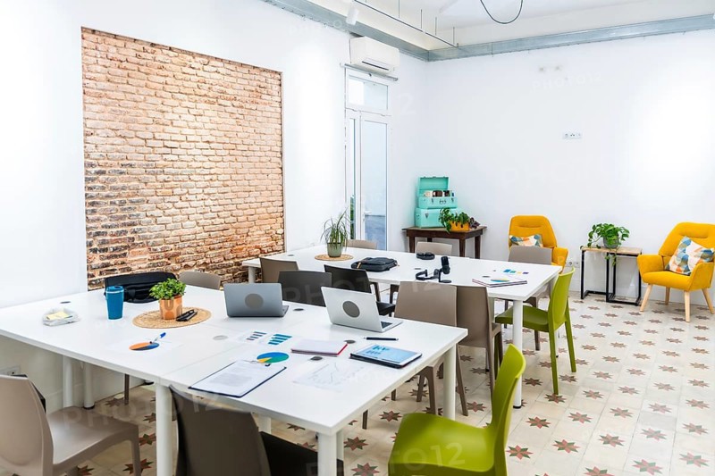 Bright open plan coworking office space featuring long white tables with laptops and charts, a textured brick wall, and comfortable yellow armchairs in a relaxed meeting corner