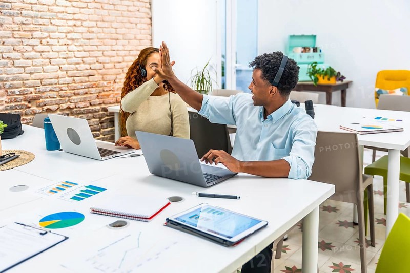 Diverse business colleagues working at laptops, high fiving to celebrate a shared achievement or successful project in a modern coworking office space