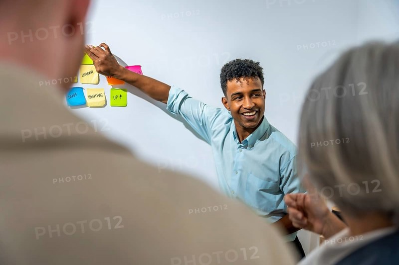 Young businessman actively leading a business strategy presentation, placing colorful sticky notes with keywords like 'leader', 'team', and 'positive' on a whiteboard for a diverse audience