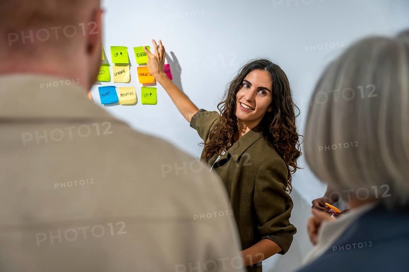Young woman presenting business concepts and brainstorming ideas to colleagues using colorful sticky notes on a white board during a corporate meeting in an office environment