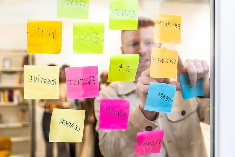 Man in a modern office space arranging colorful sticky notes with various concepts and keywords on a glass wall, actively engaged in a strategic planning and brainstorming session