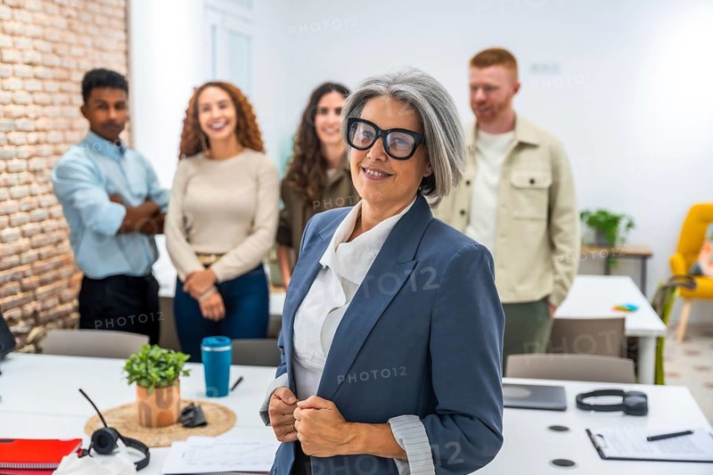 Experienced senior woman standing confidently in blue blazer and glasses, leading and collaborating with her diverse multi ethnic business team in a professional office environment