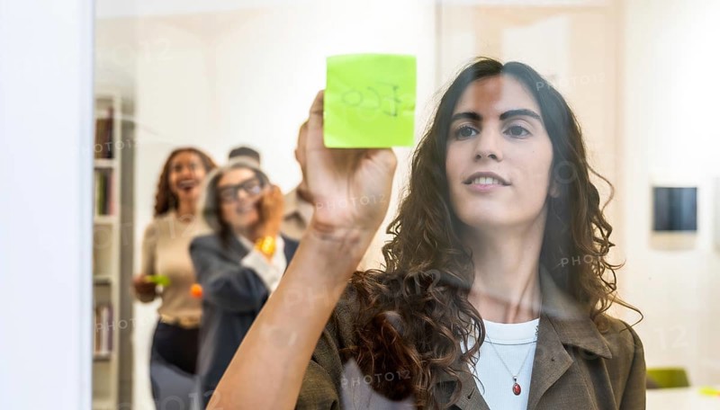 Young businesswoman sticking a green sticky note on a transparent glass wall during a team meeting, her colleagues in the background engaging in collaborative brainstorming and creative discussions