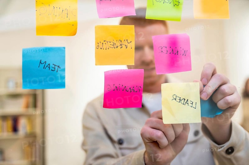 Red haired man organizing colorful sticky notes with handwritten words about planning, team, and leadership on a clear glass board during a focused brainstorming session at the office