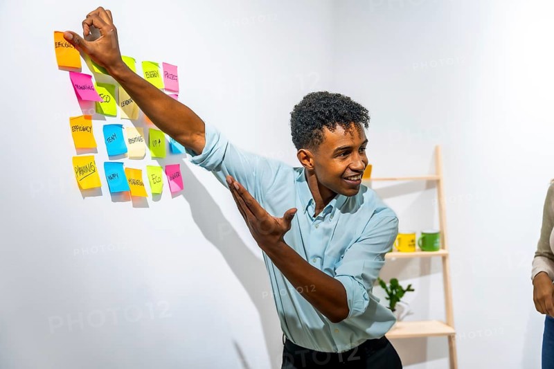 Young black man smiling and brainstorming, placing colorful sticky notes with business related words onto a white wall, organizing concepts for a project or strategy in a modern office