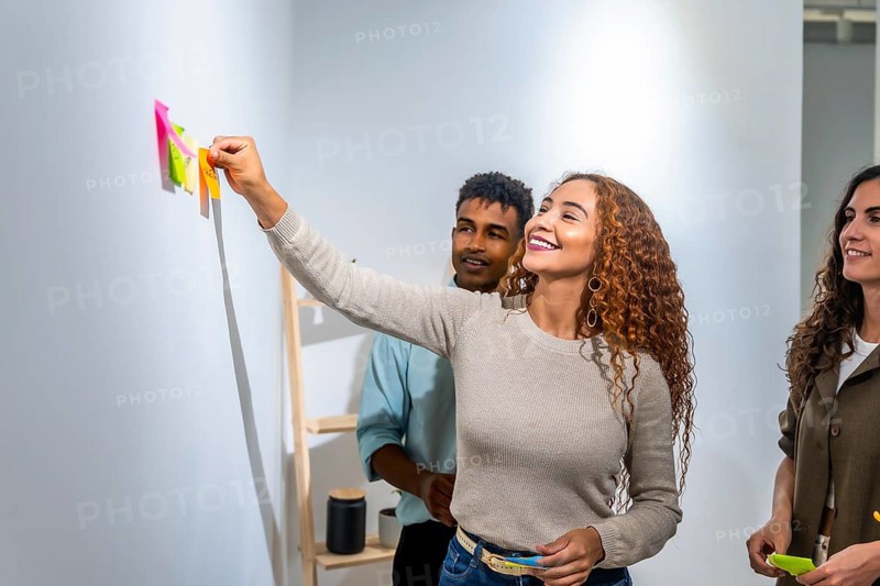 Young diverse team collaborating on a project, happy woman placing a colorful sticky note on a white wall during a brainstorming session in a modern office