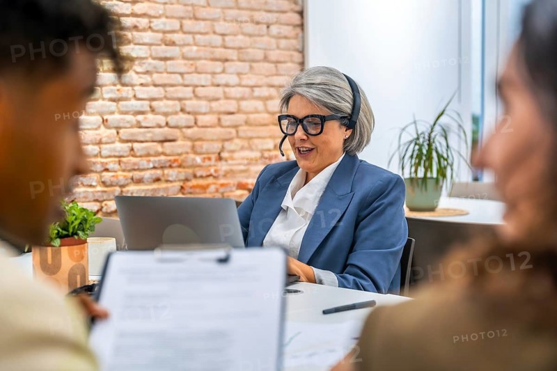 Senior businesswoman with a headset providing attentive customer service and digital support while sitting at a desk with a laptop, assisting clients in a modern office