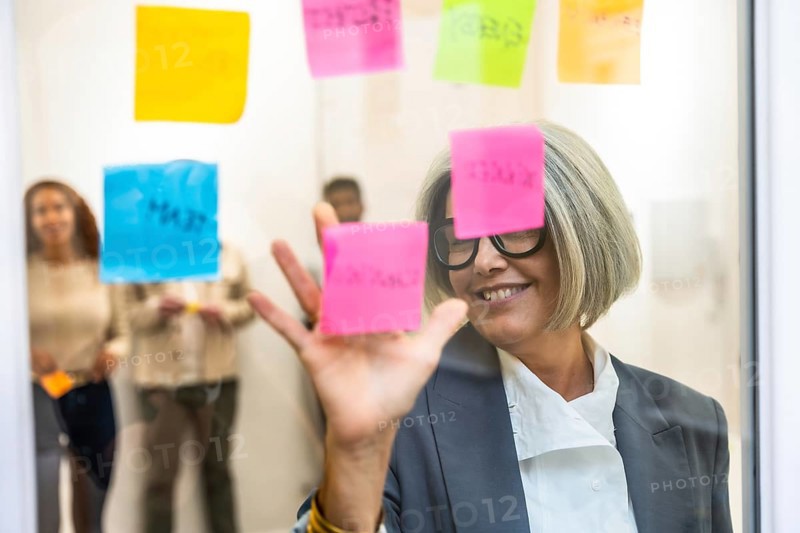 Senior business woman smiling, organizing colorful sticky notes on a glass whiteboard during a collaborative brainstorming session with colleagues in an office meeting