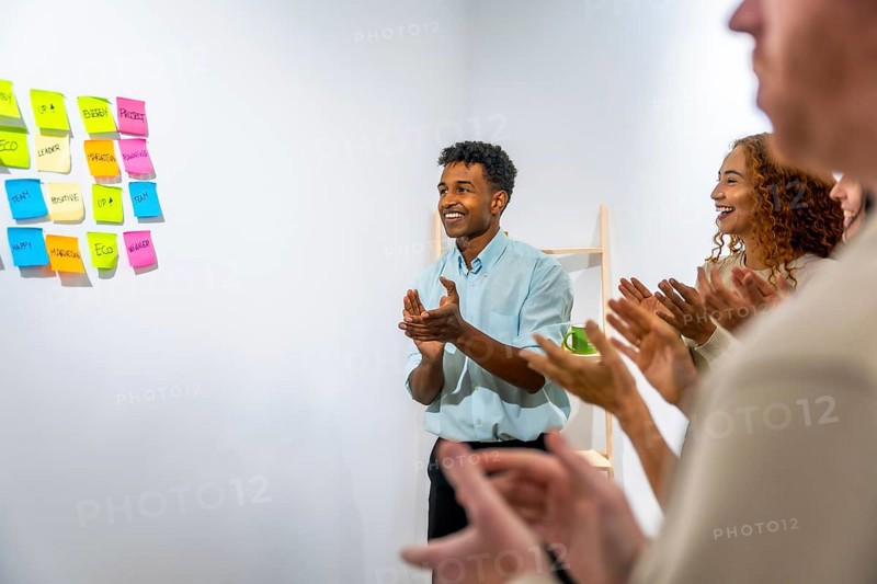 Smiling business team members are clapping, showing appreciation and recognition during a successful office meeting or presentation, with colorful sticky notes on a wall