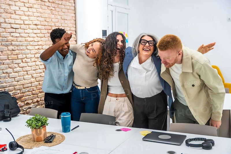 Diverse business team embracing each other excitedly, laughing and celebrating success together in a modern office meeting room, showing strong teamwork and positive corporate culture