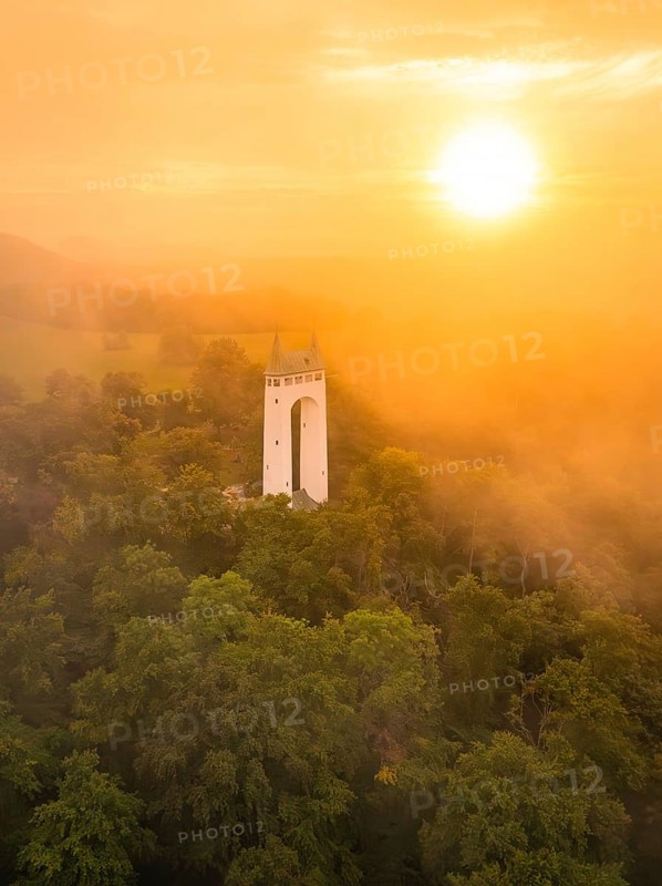 White tower rises out of misty forest while the sun illuminates orange sky, Schönbergturm, Pfullingen, Swabian Alb, Germany