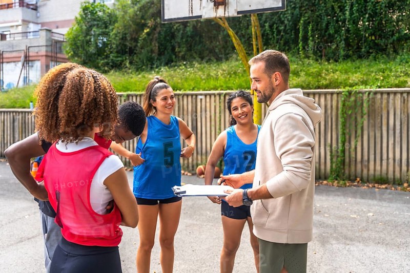 Basketball coach holding clipboard and giving instructions to a diverse group of female players during training on an outdoor court, promoting teamwork, sportsmanship, and athletic development