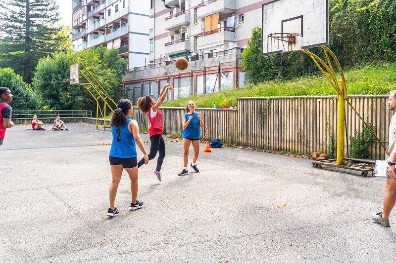 Female basketball player jumping and shooting the ball during a training session on an outdoor court, with her teammates and coach watching