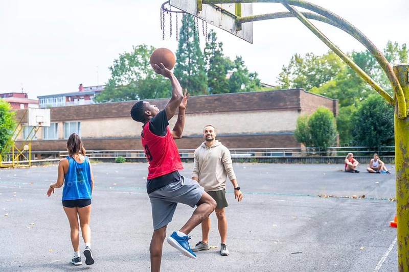 Group of friends enjoying a game of basketball on an outdoor court, with a young man shooting the ball while his friends cheer him on, embodying teamwork and camaraderie