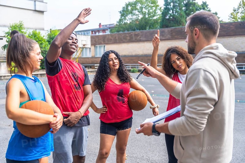Basketball coach holding a clipboard, explaining game strategy while engaging with team players during an outdoor training session on the court, fostering teamwork and skill development