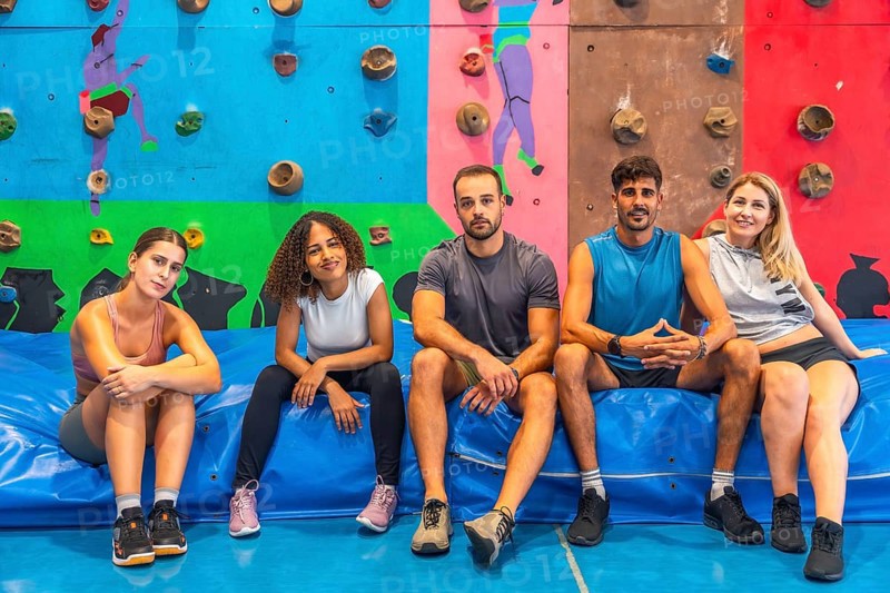 Five athletes resting on a vibrant blue crash pad in a colorful bouldering gym, enjoying a well deserved break after an intense climbing session filled with teamwork and camaraderie