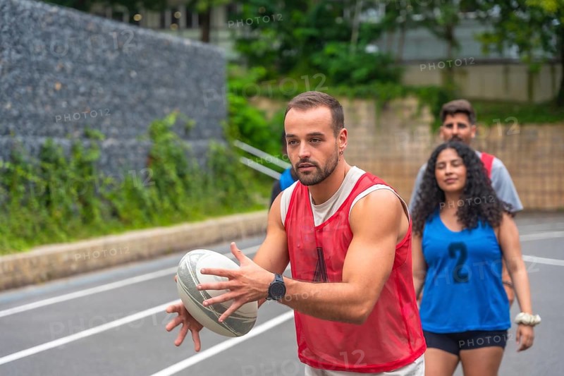 Male rugby player wearing red training bib holding and passing rugby ball on outdoor court during team practice, with teammates walking behind him