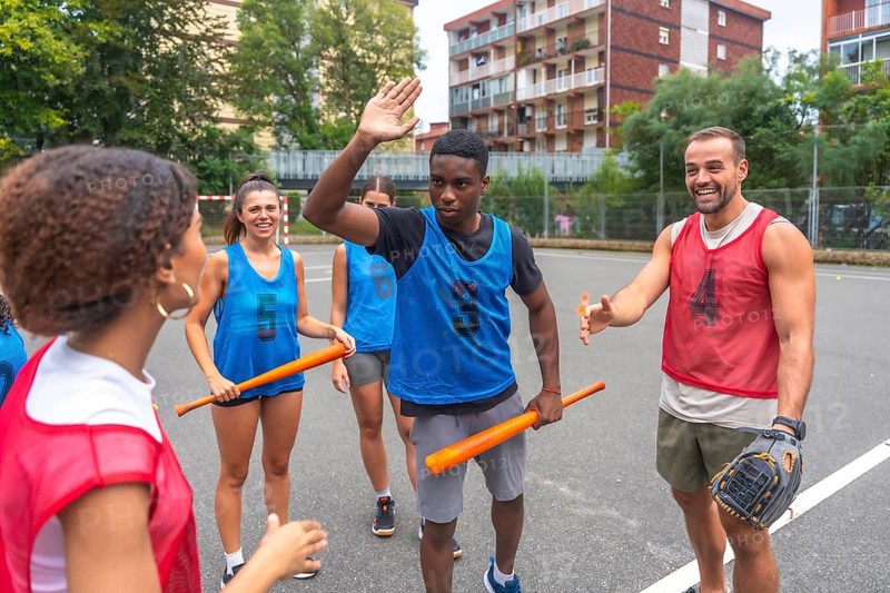 Group of cheerful multi ethnic friends wearing sportswear giving high fives and holding bats while enjoying a friendly baseball match on a sunny day on an outdoor court