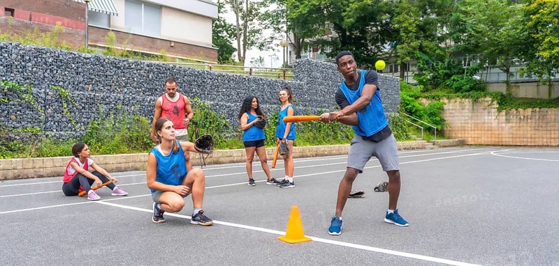 Multi ethnic baseball players practicing on an urban field, with a batter hitting the ball and a catcher poised to receive, highlighting teamwork and athleticism