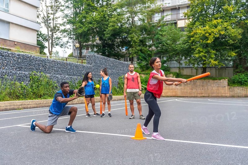 Group of friends enjoying a lively game of baseball on an outdoor court, with a woman swinging an orange bat while a male friend gets ready to catch the ball