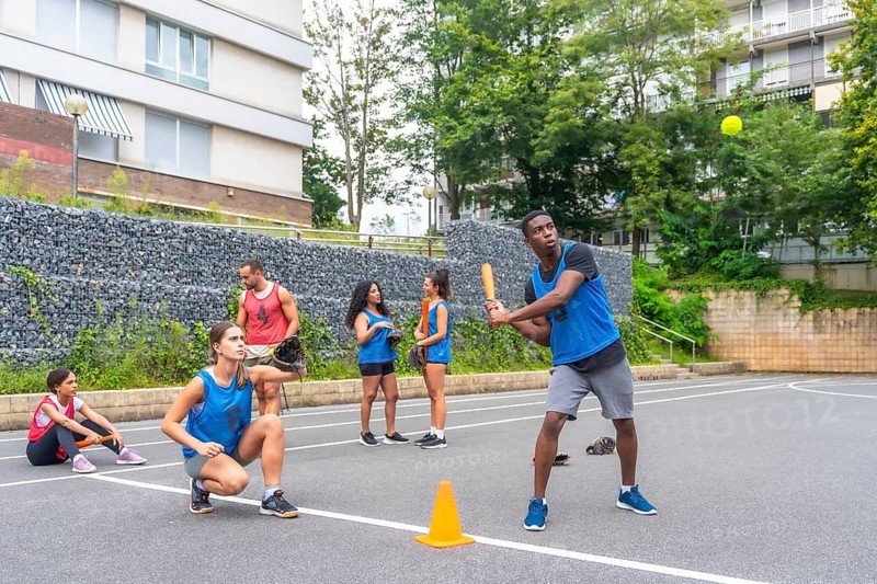 Group of young friends playing baseball in an urban environment, with a male player hitting the ball while his teammates watch and wait their turn