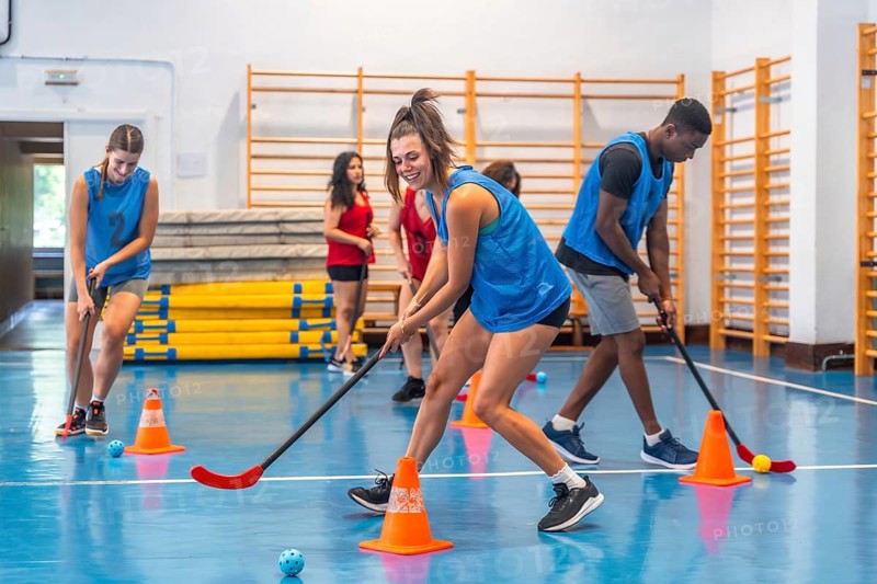 Female and male athletes wearing blue t shirts are practicing floorball, dribbling with sticks around orange cones in a gym with a blue floor and yellow equipment
