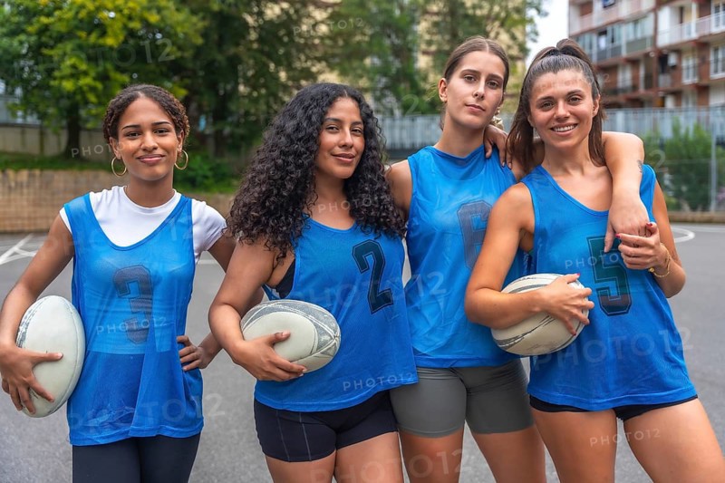 Four diverse female rugby athletes in blue jerseys stand together on an urban court after training, holding rugby balls and demonstrating camaraderie and athleticism
