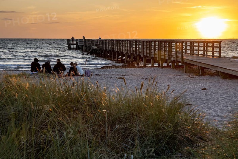Group of people sitting on the beach next to a pier at sunset, surrounded by grass and sea, Kobaek Strand, Sjaelland, Denmark