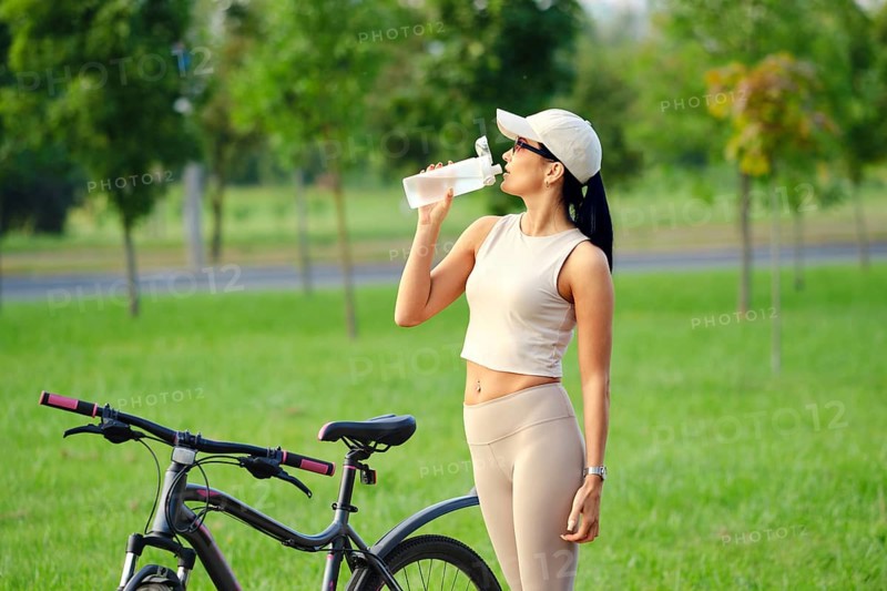 A woman dressed in activewear takes a refreshing drink from her water bottle while standing next to her bicycle in a sunny park surrounded by trees