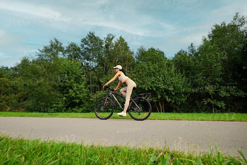 A woman rides her mountain bike on a smooth path in a lush green landscape. Tall trees line both sides of the trail, creating a tranquil atmosphere during a bright day