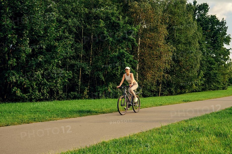 A woman rides a bicycle along a paved path surrounded by tall trees and vibrant greenery. The sunny weather creates a cheerful atmosphere for outdoor activities