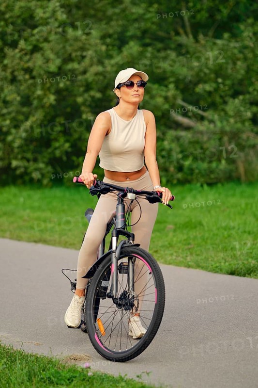 A woman enjoys a leisurely ride on her bicycle along a paved path in a vibrant green park. She wears casual athletic clothing and sunglasses while taking in the beautiful surroundings