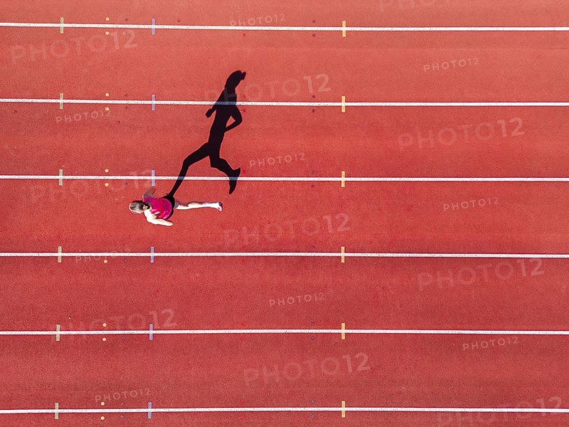 Aerial view of a female individual (runner) on a red athletics track (tartan track) with shadow as a silhouette