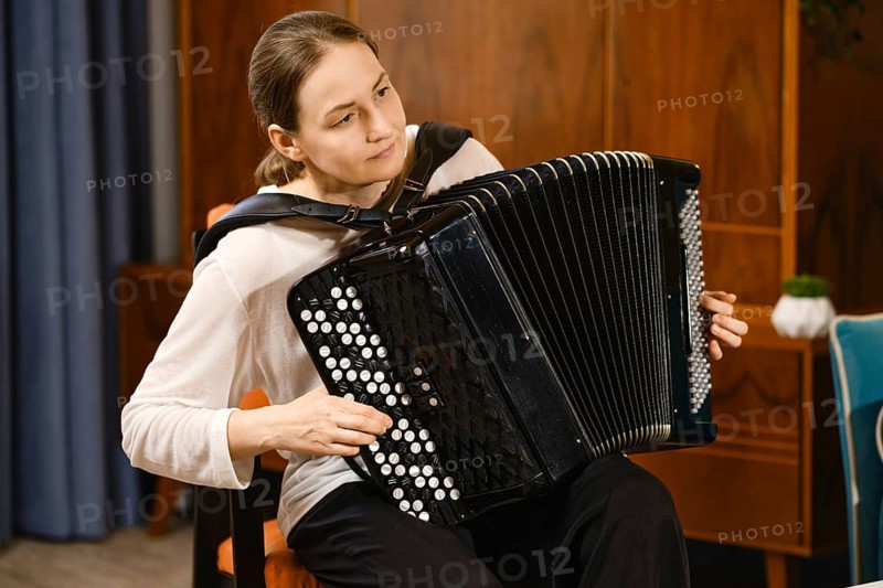 A middle-aged woman concentrates as she plays the accordion in a warmly decorated room, creating a serene atmosphere filled with music. The setting enhances the intimate vibe of her performance
