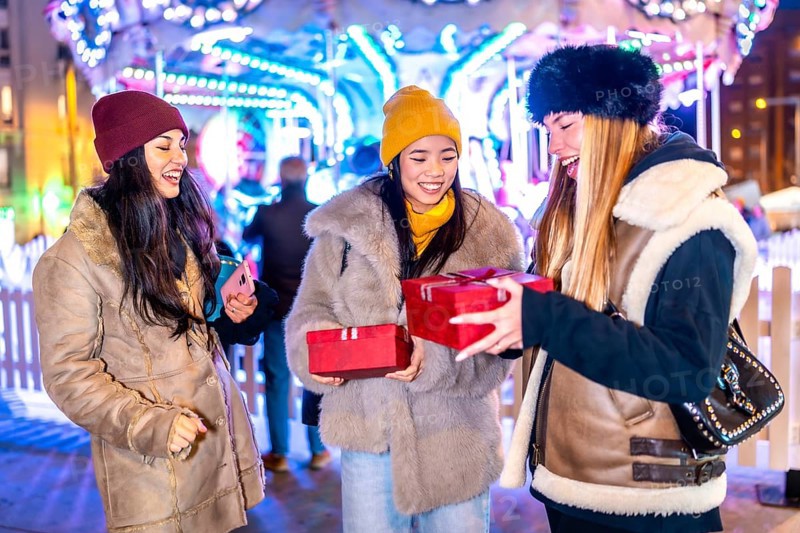 Three happy female friends exchanging christmas gifts in front of a carousel at a christmas market in the city at night