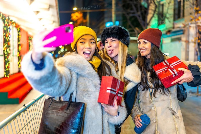 Three happy female friends taking a selfie while holding christmas gifts, enjoying a night out at a decorated christmas market
