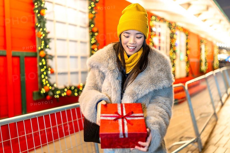 Happy young woman joyfully opening a christmas gift amidst the festive decorations of a bustling night market, surrounded by twinkling lights