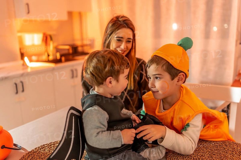 Two young brothers wearing halloween costumes playing together at home, watched by their smiling mother