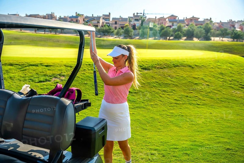 Female golfer placing her golf club into the cart while enjoying a sunny day on a lush green golf course, embracing leisure and sport