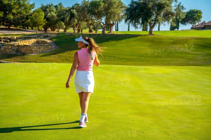 Female golfer walking away on a sunny golf course, enjoying the vibrant landscape filled with lush green grass and tall trees