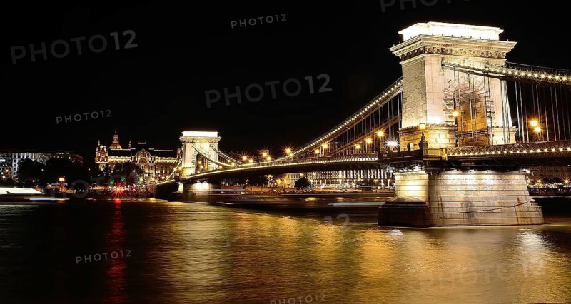 Chain bridge reflecting on danube river with gresham palace in background at night, budapest, hungary