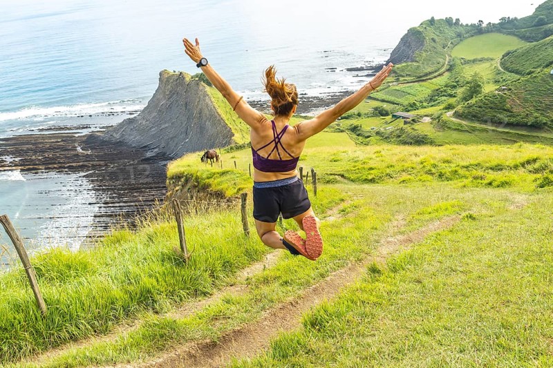 Sportswoman celebrates achieving a fitness goal by leaping in the air with arms outstretched, enjoying the breathtaking view of sakoneta beach and the flysch cliffs in zumaia, basque country