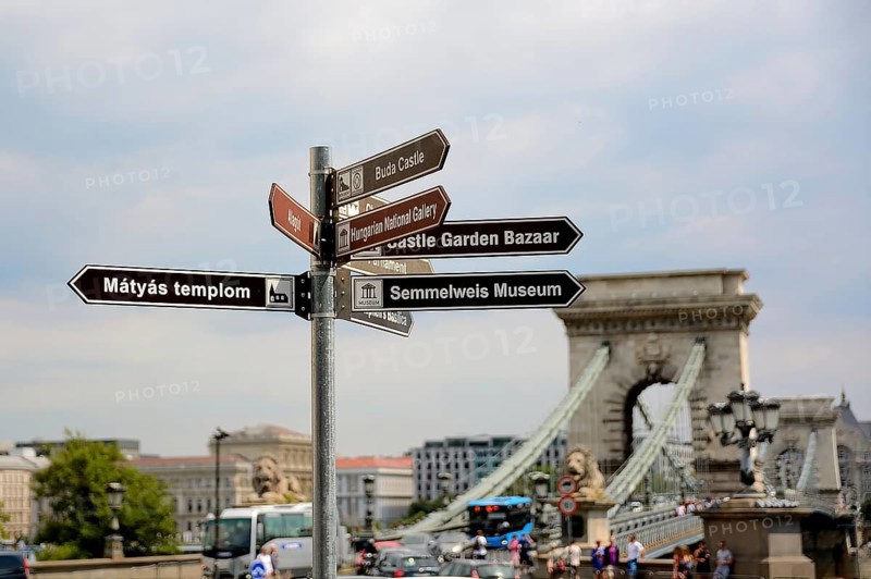 Brown tourist signs indicating directions to matthias church, buda castle, hungarian national gallery, castle garden bazaar and semmelweis museum, with chain bridge in the background