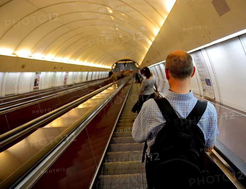Tourist with a backpack ascending an escalator in a bustling subway station, surrounded by urban architecture and modern transportation
