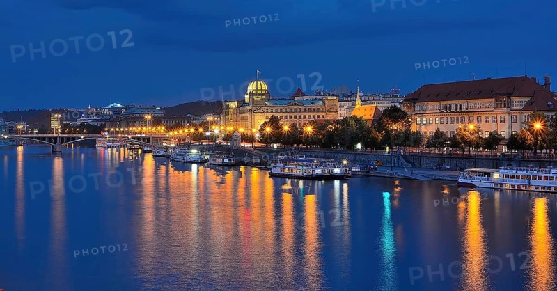 City lights reflecting in vltava river with illuminated buildings and boats moored along the embankment at dusk in prague