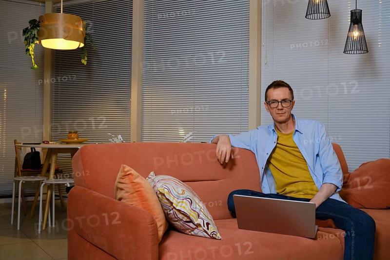 A man sits comfortably on a coral sofa, focused on his laptop in a cozy living room decorated with warm lighting. He is watching a new episode of the series online