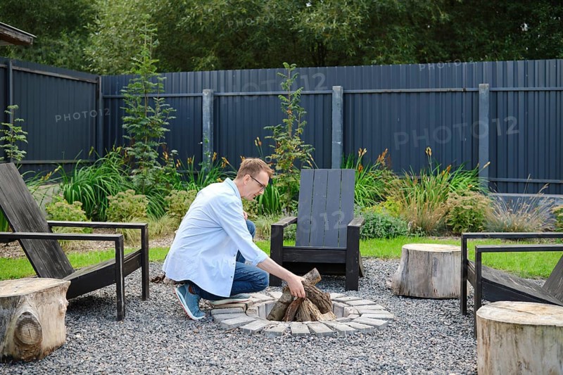 A middle-aged man dressed in a light blue shirt arranges firewood at a circular stone fire pit surrounded by wooden seating